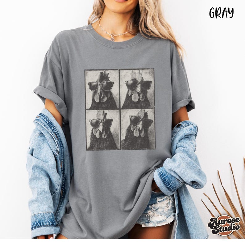 Op de afbeelding: Grijze t-shirt met een vierpanelen grafische afbeelding van kippen met zonnebrillen. Het shirt heeft een relaxte pasvorm en opgerolde mouwen. Het woord "Gray" staat rechtsboven gedrukt. De merknaam "Aurose Studio" staat rechtsonder gedrukt.