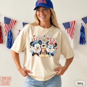 May include: Ivory-colored t-shirt featuring a graphic of Mickey Mouse, Pluto, and Minnie Mouse in patriotic attire. The design includes fireworks and stars. The model is wearing a blue baseball cap with "USA" on it. The shirt is from Comfort Colors.