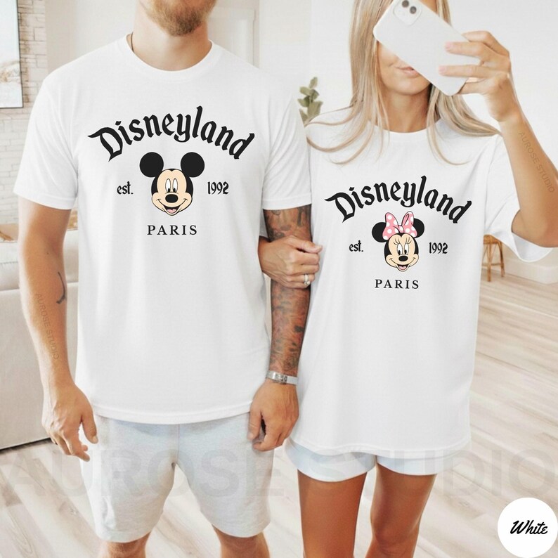 May include: Two white t-shirts with the Disneyland Paris logo. One shirt features Mickey Mouse, the other Minnie Mouse with a pink bow. The text "Disneyland est. 1992 PARIS" is printed in black script.