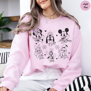 May include: Light pink sweatshirt featuring black and white illustrations of Disney characters, including Mickey Mouse, Minnie Mouse, Pluto, Daisy Duck, Goofy, and Donald Duck. The sweatshirt has a crew neck and long sleeves.