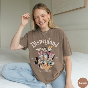 May include: Brown t-shirt with the word "Disneyland" in a vintage font, along with cartoon characters. The shirt also features the text "est. 1955" and "CALIFORNIA".