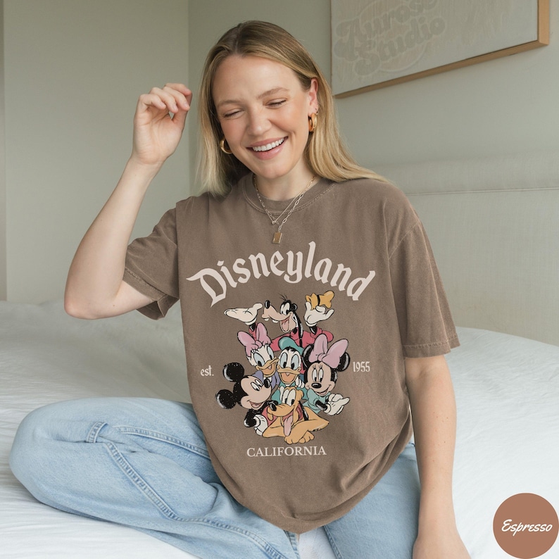 May include: Brown t-shirt with the word "Disneyland" in a vintage font, along with cartoon characters. The shirt also features the text "est. 1955" and "CALIFORNIA".