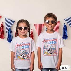 May include: Two children wearing white t-shirts with a colorful graphic celebrating the United States of America's 250th Anniversary. The design includes cartoon characters, fireworks, and the text "Anniversary 250 Years of Freedom."