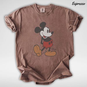 May include: Espresso-colored t-shirt featuring a classic Mickey Mouse graphic. The cartoon character is depicted in a walking pose, wearing red shorts, yellow shoes, and black gloves. The shirt has a crew neck and short sleeves.