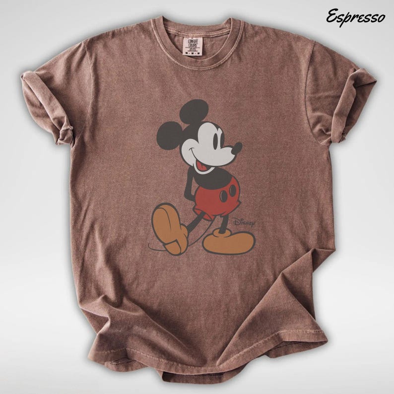 May include: Espresso-colored t-shirt featuring a classic Mickey Mouse graphic. The cartoon character is depicted in a walking pose, wearing red shorts, yellow shoes, and black gloves. The shirt has a crew neck and short sleeves.