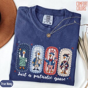 May include: A navy blue Comfort Colors t-shirt featuring a patriotic goose design. The graphic shows four geese dressed as American icons, with the text "Just a patriotic goose." A gold necklace is on the shirt.