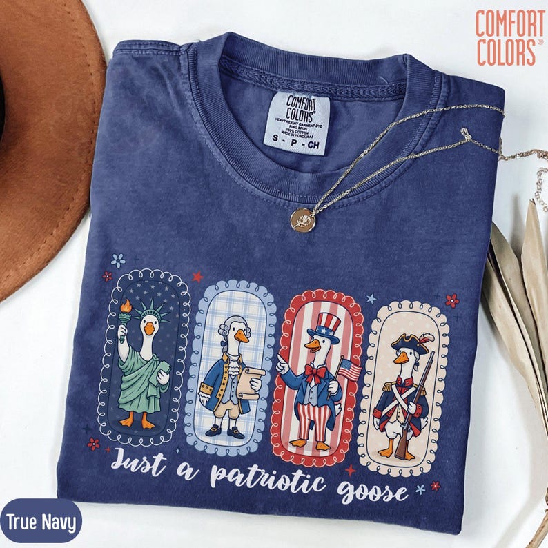 May include: A navy blue Comfort Colors t-shirt featuring a patriotic goose design. The graphic shows four geese dressed as American icons, with the text "Just a patriotic goose." A gold necklace is on the shirt.