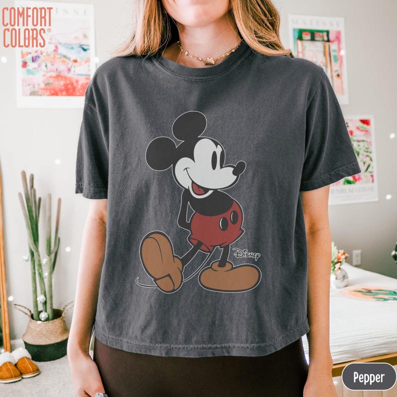May include: Dark gray Comfort Colors t-shirt featuring a classic Mickey Mouse graphic. The cartoon character is depicted in black, white, and red, with the Disney logo. The shirt has short sleeves and a crew neck.