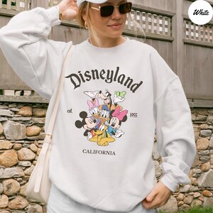 K&ouml;nnte beinhalten: Wei&szlig;es Sweatshirt mit dem Wort "Disneyland" in einer stilisierten Schriftart und dem Text "est. 1955" darunter. Eine farbenfrohe Grafik von Disney-Figuren ist unter dem Text zentriert, mit dem Wort "CALIFORNIA" unten.