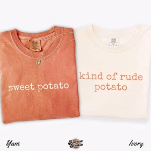 May include: Two t-shirts, one yam-colored and one ivory, with the phrases "sweet potato" and "kind of rude potato" printed on them. A gold necklace with a pendant rests on the yam-colored shirt. The shirts are from the Comfort Colors brand.