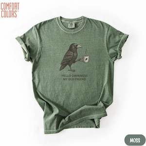 May include: A moss green crew neck t-shirt featuring a graphic illustration of a black crow holding a steaming mug. Below the crow, the text reads "HELLO DARKNESS MY OLD FRIEND". The shirt has a vintage-style washed appearance and short sleeves.