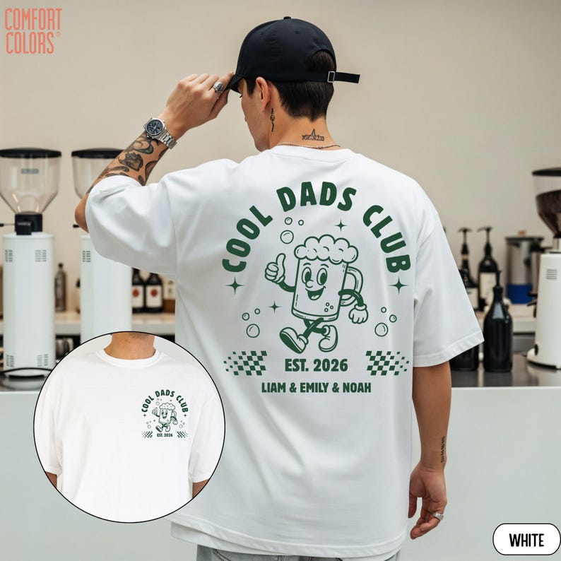 以下が含まれることがあります： 緑色の「COOL DADS CLUB」のグラフィックが特徴の白いTシャツ。親指を立てている漫画のビールジョッキが描かれています。「EST. 2026」の文字と「LIAM & EMILY & NOAH」の名前もプリントされています。 Comfort Colorsブランドです。