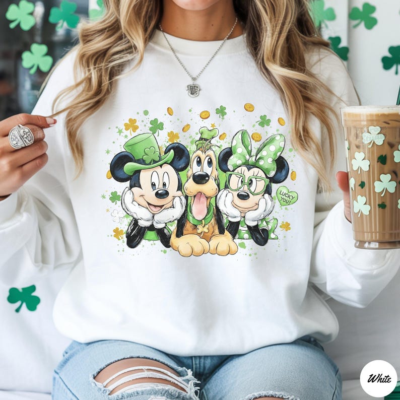 May include: White sweatshirt featuring Mickey Mouse, Goofy, and Minnie Mouse in St. Patrick's Day attire. Mickey wears a green top hat, Goofy has a green shamrock, and Minnie has a green bow and glasses. The phrase "Lucky You!" is on a green heart.