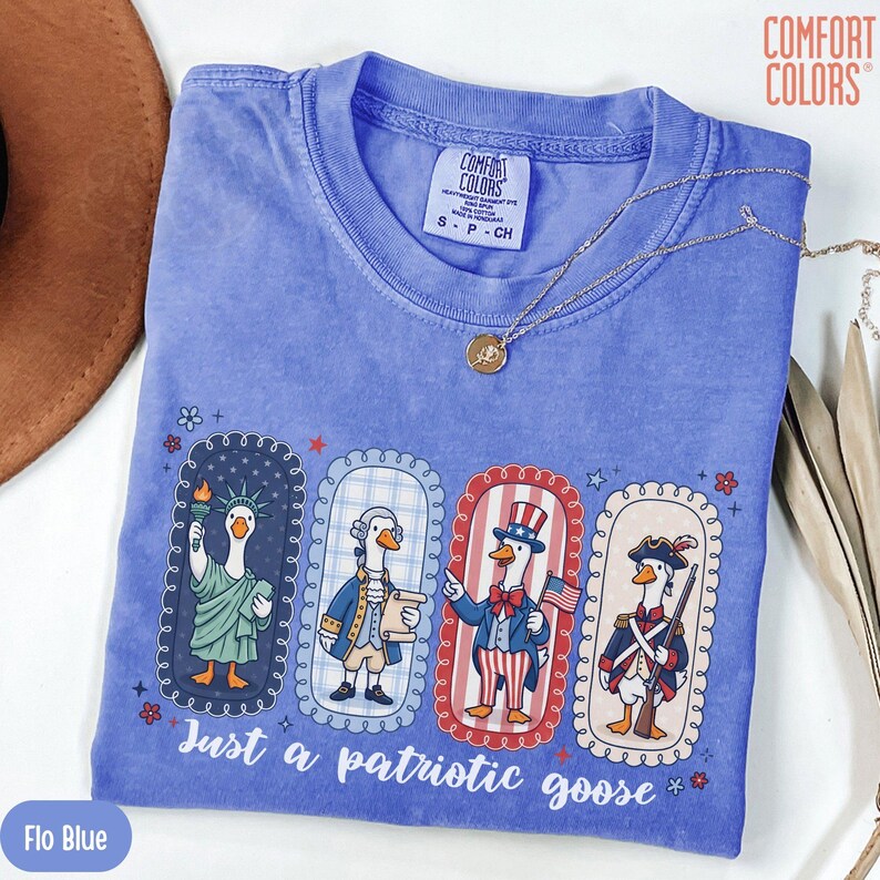 May include: Flo Blue Comfort Colors t-shirt with a patriotic goose design. The design features four geese dressed in patriotic attire, with the text "Just a patriotic goose". A gold necklace is draped across the shirt.