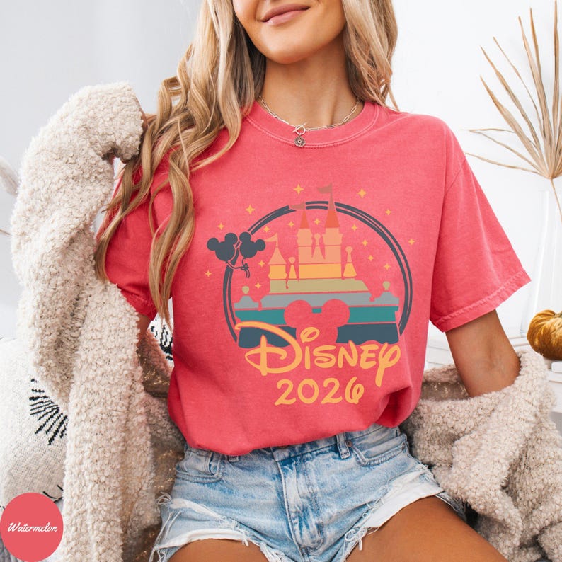 May include: A red t-shirt featuring a colorful graphic of a castle, rainbow, and the text "Disney 2026". The design is enclosed in a circular frame with Mickey Mouse balloons. The shirt is worn with denim shorts.