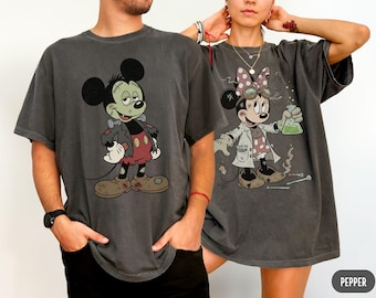 May include: Two charcoal gray t-shirts featuring cartoon character graphics. The left shirt displays a green-skinned character with stitches and a dark jacket. The right shirt shows a character in a lab coat holding a glowing green flask.