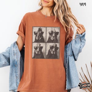 Op de afbeelding: Roestkleurig T-shirt met een vierpaneelsafbeelding van een haan met een zonnebril. Het shirt heeft korte mouwen en een relaxte pasvorm. De tekst "YAM" staat rechtsboven gedrukt. Het merklogo "Aurose Studio" staat rechtsonder gedrukt.