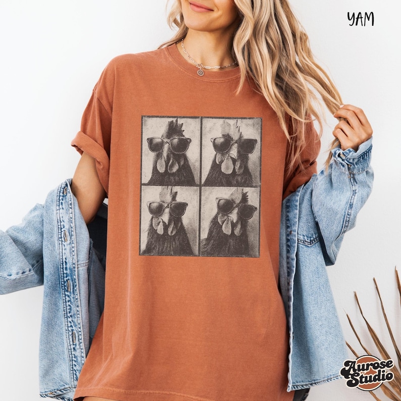 Op de afbeelding: Roestkleurig T-shirt met een vierpaneelsafbeelding van een haan met een zonnebril. Het shirt heeft korte mouwen en een relaxte pasvorm. De tekst "YAM" staat rechtsboven gedrukt. Het merklogo "Aurose Studio" staat rechtsonder gedrukt.