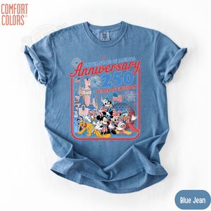 May include: Blue jean Comfort Colors t-shirt featuring a graphic celebrating the 250th Anniversary of the United States of America. The graphic includes cartoon characters, fireworks, and the text "Anniversary 250 Years of Freedom."