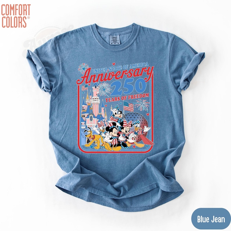 May include: Blue jean Comfort Colors t-shirt featuring a graphic celebrating the 250th Anniversary of the United States of America. The graphic includes cartoon characters, fireworks, and the text "Anniversary 250 Years of Freedom."
