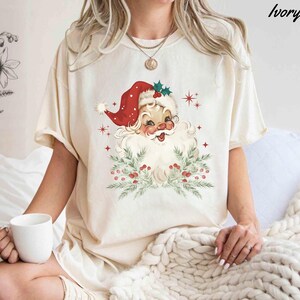 May include: Ivory-colored t-shirt featuring a vintage Santa Claus graphic. Santa is depicted with a red hat, white beard, and a wreath of greenery and berries. The shirt has a relaxed fit and short sleeves, perfect for the holiday season.