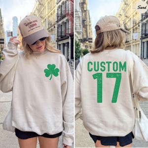 K&ouml;nnte beinhalten: Ein sandfarbenes Sweatshirt mit einem gr&uuml;nen Kleeblatt auf der Vorderseite und dem Wort "CUSTOM" &uuml;ber der Zahl "17" auf dem R&uuml;cken. Die Person tr&auml;gt eine beige Baseballkappe mit Text.