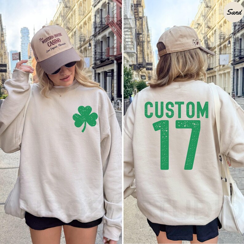 K&ouml;nnte beinhalten: Ein sandfarbenes Sweatshirt mit einem gr&uuml;nen Kleeblatt auf der Vorderseite und dem Wort "CUSTOM" &uuml;ber der Zahl "17" auf dem R&uuml;cken. Die Person tr&auml;gt eine beige Baseballkappe mit Text.