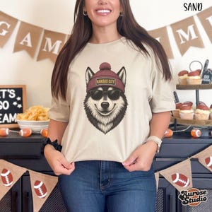 Op de afbeelding: Een zandkleurig T-shirt met een afbeelding van een husky met een zonnebril en een bordeauxrode muts met "Kansas City" erop. De hondenafbeelding is zwart, wit en bruin. Het shirt wordt gecombineerd met een blauwe jeans.