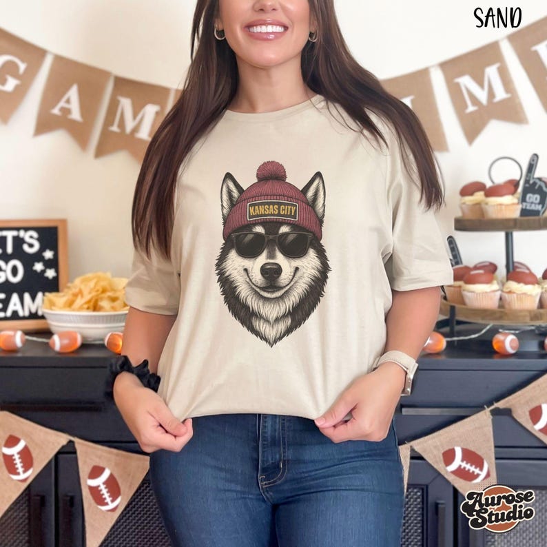 Op de afbeelding: Een zandkleurig T-shirt met een afbeelding van een husky met een zonnebril en een bordeauxrode muts met "Kansas City" erop. De hondenafbeelding is zwart, wit en bruin. Het shirt wordt gecombineerd met een blauwe jeans.