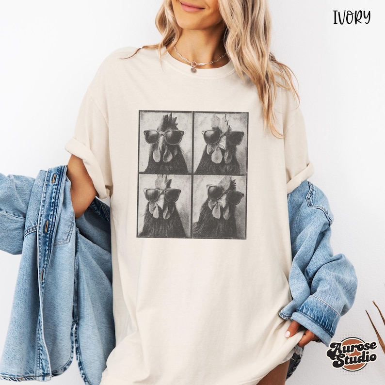Op de afbeelding: Ivoor kleurig T-shirt met een vierpaneels afbeelding van een haan met zonnebril. Het shirt heeft een relaxte pasvorm en opgerolde mouwen. De tekst "Ivory" staat rechtsboven. Het Aurose Studio logo staat rechtsonder.