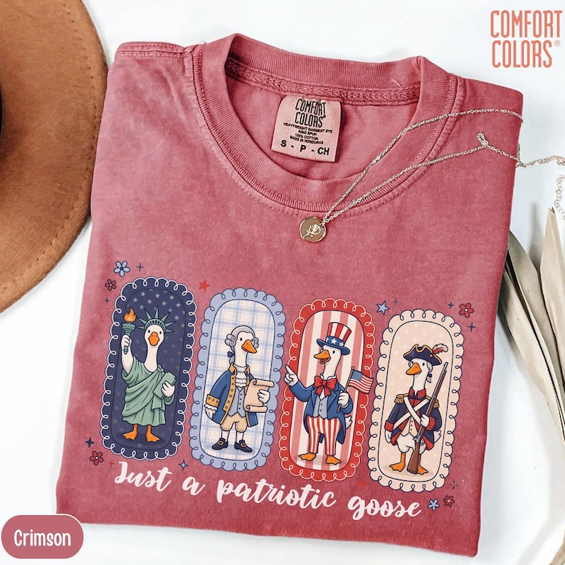 May include: Crimson t-shirt featuring a graphic of geese in patriotic outfits. The design includes geese dressed as historical figures, with the text "Just a patriotic goose". A silver necklace with a pendant is on the shirt.