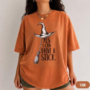 Puede incluir: Una camiseta de manga corta color naranja quemado con un gr&aacute;fico negro de un sombrero de bruja sobre una escoba. El texto dice YES I CAN DRIVE A STICK. La prenda tiene cuello redondo y un corte holgado con un estampado oscuro y llamativo.