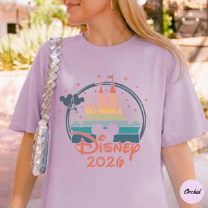 May include: A light purple t-shirt featuring a colorful Disney castle design within a circular frame. The design includes the text "Disney 2026" in a script font. The shirt is a casual style.