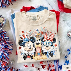 May include: Ivory-colored t-shirt featuring Mickey Mouse, Pluto, and Minnie Mouse in patriotic attire. The graphic includes fireworks, hearts, and stars. Additional shirts in red, white, and blue are visible in the background. The t-shirt is labeled "Comfort Colors".