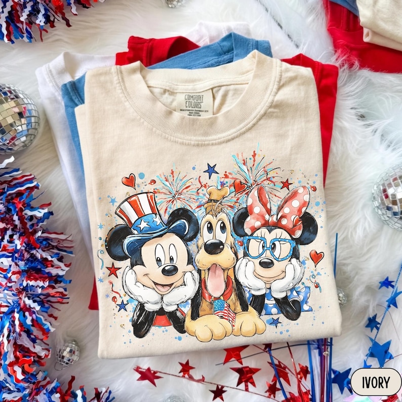 May include: Ivory-colored t-shirt featuring Mickey Mouse, Pluto, and Minnie Mouse in patriotic attire. The graphic includes fireworks, hearts, and stars. Additional shirts in red, white, and blue are visible in the background. The t-shirt is labeled "Comfort Colors".