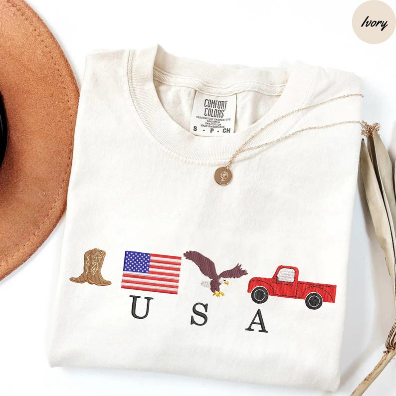 May include: Ivory-colored t-shirt with embroidered patriotic symbols: a cowboy boot, American flag, eagle, and red pickup truck. The word "USA" is embroidered below the symbols. A gold necklace with a pendant is draped across the shirt.