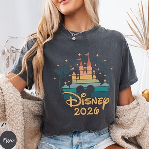 May include: Dark gray t-shirt featuring a colorful graphic of a castle, Mickey Mouse ears, and the text "Disney 2026." The design includes shades of orange, yellow, green, and blue. The shirt has a vintage, faded look.
