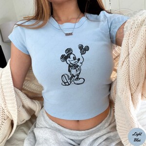 Op de afbeelding: Lichtblauw crop top met een zwarte schets van Mickey Mouse met ballonnen. De top heeft korte mouwen en een ronde halslijn. De tekst "Light Blue" staat rechtsonder.