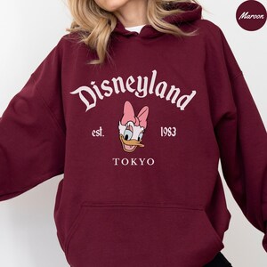 May include: Maroon hoodie with a hood featuring the Disneyland logo in white script, Daisy Duck graphic, and the text "est. 1983 TOKYO". The sweatshirt is a solid maroon color.