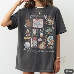 K&ouml;nnte beinhalten: Dunkelgraues T-Shirt mit einer farbenfrohen Collage von Disney-Figuren und Wahrzeichen. Das Design zeigt Micky Maus, Minnie Maus, Donald Duck und Chip und Chap, mit dem Text "Walt Disneyland" und "2026".