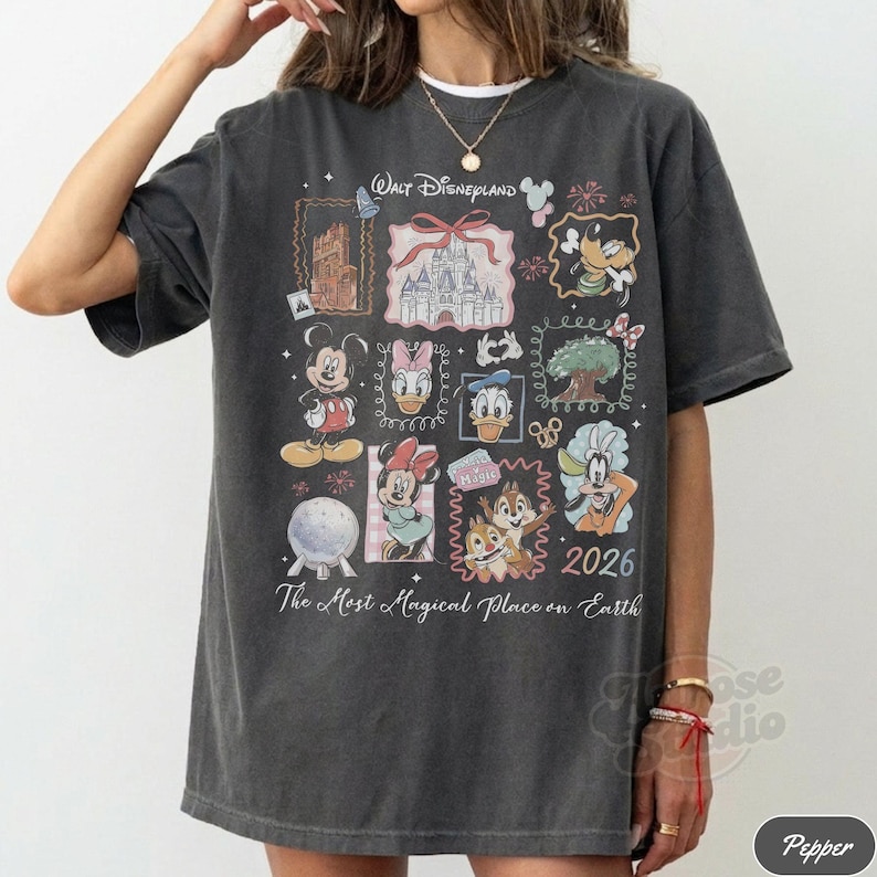 K&ouml;nnte beinhalten: Dunkelgraues T-Shirt mit einer farbenfrohen Collage von Disney-Figuren und Wahrzeichen. Das Design zeigt Micky Maus, Minnie Maus, Donald Duck und Chip und Chap, mit dem Text "Walt Disneyland" und "2026".