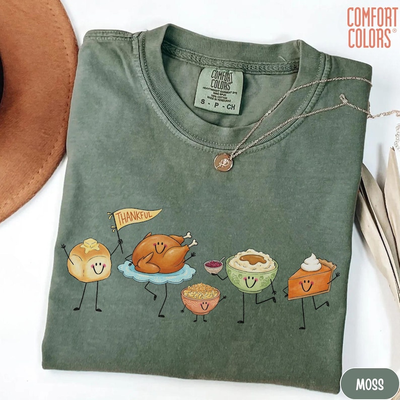 May include: A moss green short-sleeve t-shirt featuring a graphic of anthropomorphic Thanksgiving foods including a dinner roll with a flag reading Thankful, a roasted turkey, stuffing, mashed potatoes, and a slice of pumpkin pie with whipped cream.
