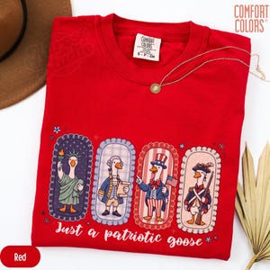 May include: A red t-shirt featuring a patriotic goose design. The graphic shows four geese in historical and patriotic outfits, with the text "Just a patriotic goose." A gold necklace is draped across the shirt.