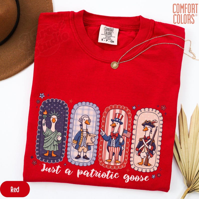 May include: A red t-shirt featuring a patriotic goose design. The graphic shows four geese in historical and patriotic outfits, with the text "Just a patriotic goose." A gold necklace is draped across the shirt.
