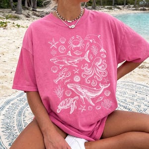 Pode incluir: Uma t-shirt de manga curta em cor-de-rosa brilhante com uma impress&atilde;o gr&aacute;fica branca de v&aacute;rias criaturas marinhas, incluindo um tubar&atilde;o, uma baleia, um polvo, uma tartaruga marinha, um caranguejo, uma lagosta, um golfinho, estrelas-do-mar, corais e conchas.