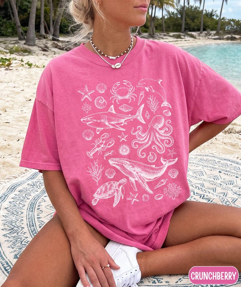 Pode incluir: Uma t-shirt de manga curta em cor-de-rosa brilhante com uma impress&atilde;o gr&aacute;fica branca de v&aacute;rias criaturas marinhas, incluindo um tubar&atilde;o, uma baleia, um polvo, uma tartaruga marinha, um caranguejo, uma lagosta, um golfinho, estrelas-do-mar, corais e conchas.