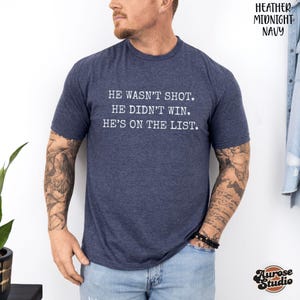 Może przedstawiać: Granatowa koszulka z napisem "HE WASN'T SHOT. HE DIDN'T WIN. HE'S ON THE LIST." Koszulka jest w kolorze granatowym i ma kr&oacute;tkie rękawy. Logo Aurose Studio znajduje się w prawym dolnym rogu.