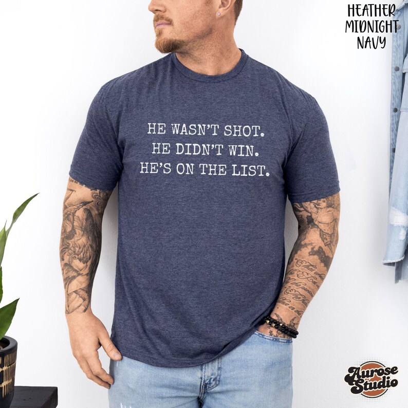 Może przedstawiać: Granatowa koszulka z napisem "HE WASN'T SHOT. HE DIDN'T WIN. HE'S ON THE LIST." Koszulka jest w kolorze granatowym i ma kr&oacute;tkie rękawy. Logo Aurose Studio znajduje się w prawym dolnym rogu.