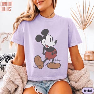 May include: A short-sleeved, orchid-colored t-shirt featuring a graphic of Mickey Mouse. The cartoon character is depicted in his classic pose, with a white face, black ears, red shorts, and brown shoes. The shirt has the "Comfort Colors" logo.