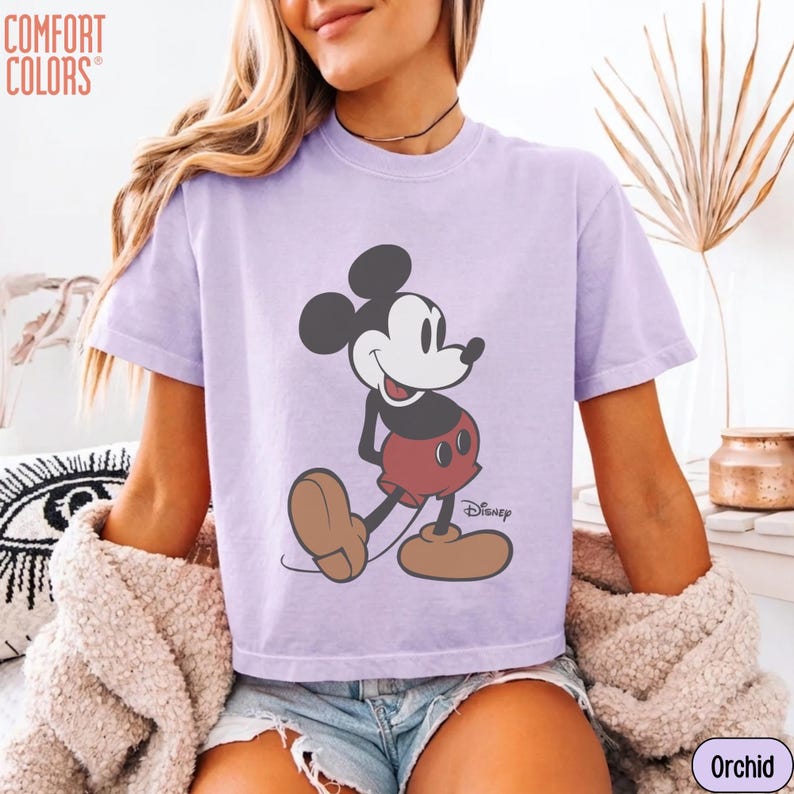 May include: A short-sleeved, orchid-colored t-shirt featuring a graphic of Mickey Mouse. The cartoon character is depicted in his classic pose, with a white face, black ears, red shorts, and brown shoes. The shirt has the "Comfort Colors" logo.